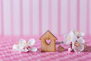 Closeup wooden house with hole in form of heart surrounded by white flowering tree branches on pink striped background.
