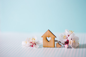 Closeup wooden house with hole in form of heart surrounded by white flowering tree branches on pastel blue background.