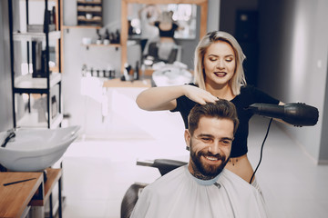 handsome young bearded guy sitting in an armchair in a beauty salon and the girl around him dry his...