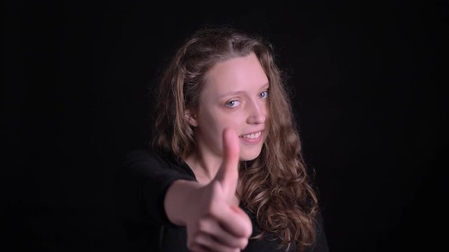 Portrait of young curly-haired girl gesturing finger-up showing like and respect joyfully into camera on black background.