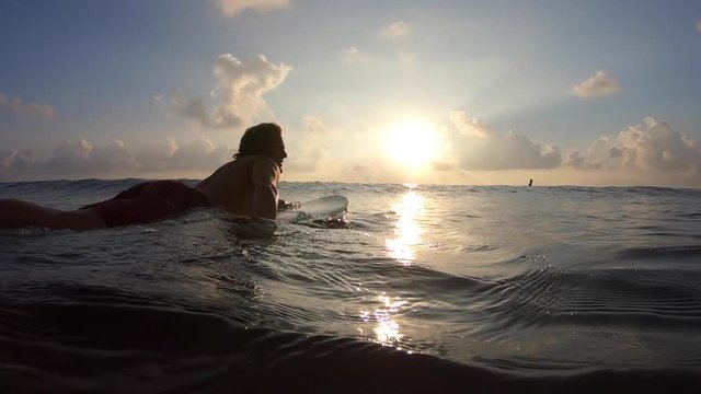 Young causacian surfer with long hairs paddles on the line up at sunrise