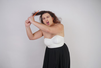 Naklejka premium emotional unhappy woman with disheveled hair tries to comb them and suffers. curvy girl stands on white background in Studio alone.