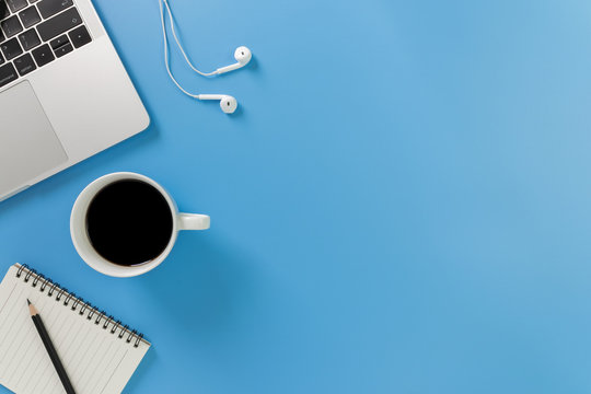 Listening to music at work. Flat lay desk top with laptop, earphone, coffee on blue background. Cool tone.