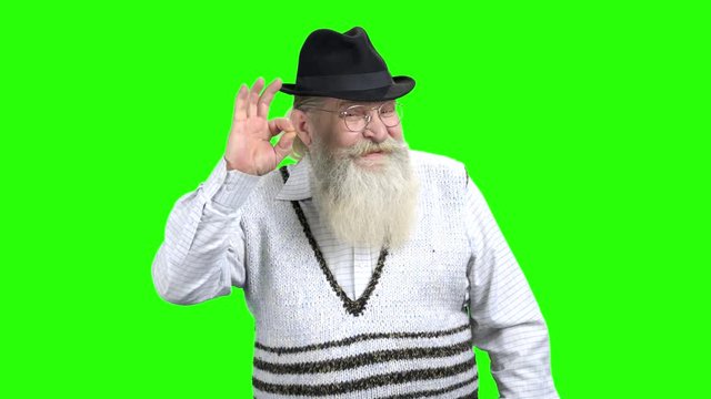 Elderly man with beard showing OK sign. Portrait of cheerful caucasian pensioner in hat on green Chroma Key background.