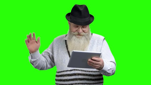 Smiling senior man holding computer tablet. Positive senior man waving with hand while holding portable tablet on green screen. Communication via internet.