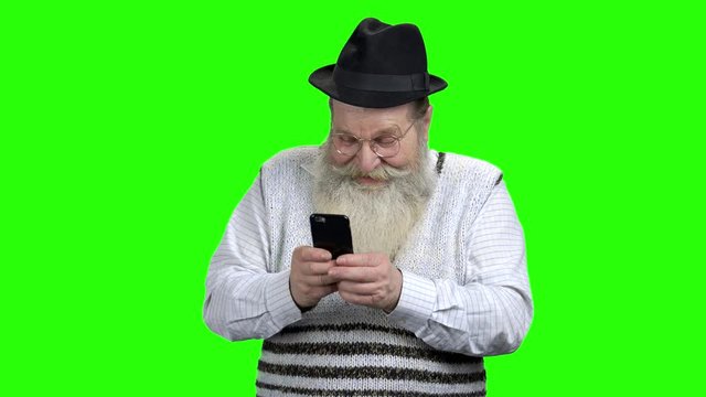 Funny caucasian pensioner using mobile phone. Old bearded man playing game on touch screen phone. Green Chroma Key background.