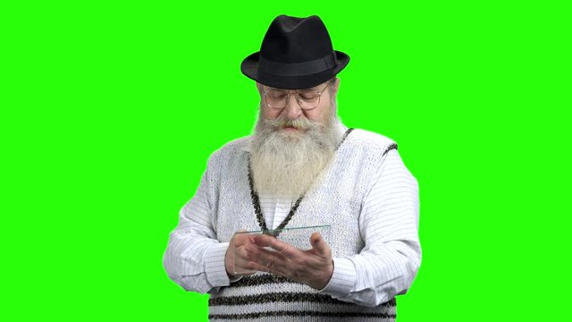 Senior man with beard using transparent plastic device. Aged man wearing hat and eyeglasses using glass digital tablet. Green screen background.