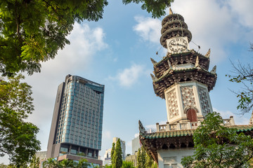 Low angle of beautiful ancient asian clock tower in city center park with branch have fresh green leaf foreground and commercial building with blue sky background