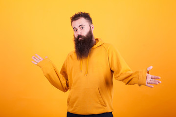 Handsome bearded man with orange hoodie looking confused over yellow background