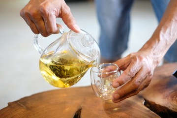 Hand that pours chinese green tea from glass kettle to small cup.