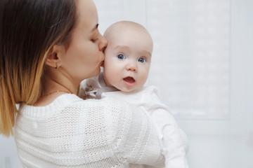 Young happy loving mother plays kisses her baby daughter in her arms against the window. Maternal care. Childcare. Close-up.