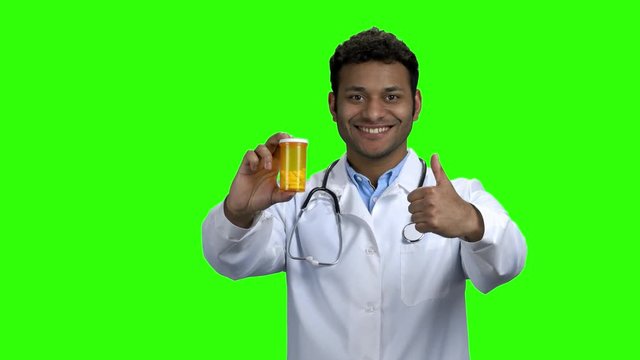 Happy doctor showing pills and thumb up. Young dark-skinned doctor holding medicaments and giving approval sign, green screen.