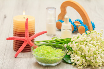 Fototapeta na wymiar Bottles with cream for face skin and aromatic oil, bowl with sea salt, burning candle, starfish, hand massager and bouquet of lilies of the valley.