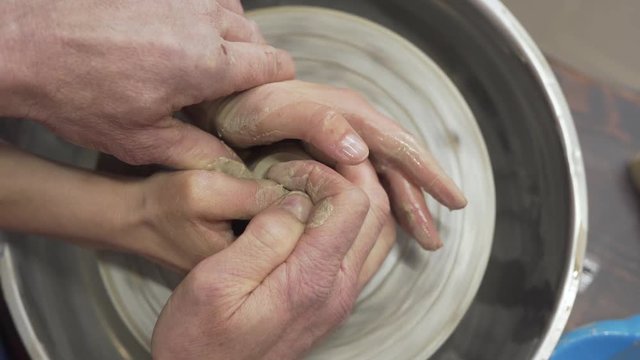 A close up video of two hands: one is younger and the other is older. Both are with cla on the spinning wheel.  A craftsman helps a student to create a ceramic vase.  The background is blurred