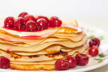 A stack of thin pancakes with red cherry berries and syrup on a plate on a white background in warm colors.
