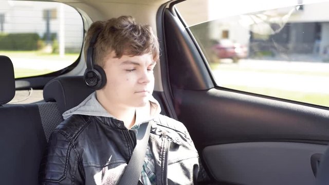 Teen boy with headphones playing game with digital tablet while sitting in the back seat of car. Happy child enjoying leisure with tablet pc in automobile during sunny day road trip. 