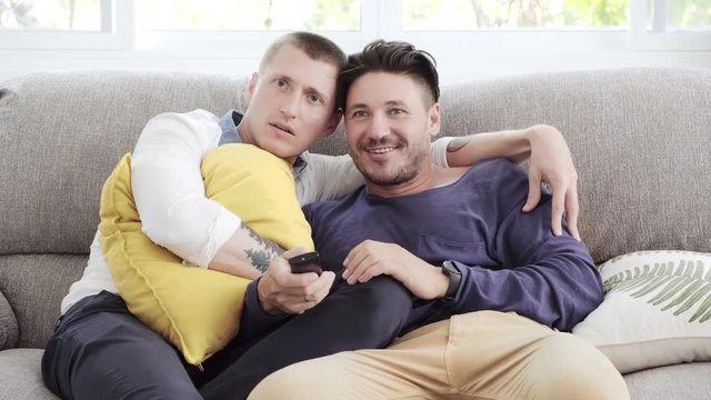 Young gay male couple relaxing on couch in living room. Caucasian, european men sitting together on sofa. Enjoy watching tv, scare mood. Happy gay friendship, relationship, lifestyle concept. 