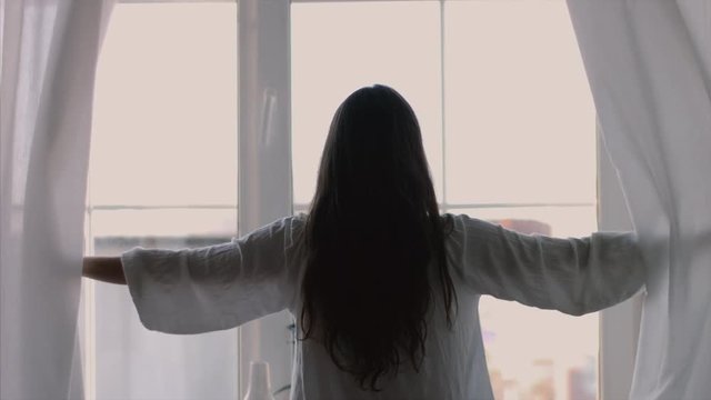 Longhaired brunette girl opening curtains window stretching in morning natural light 4k back view. Sexy woman stands raising arms hands up silhouetted indoors 4k. Freedom femininity fashion clothes
