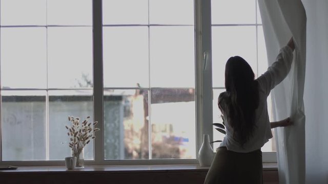 Girl pulling curtains to look out of window enjoying morning sun light back view 4k handheld camera. Natural long-haired woman stretching by window backlit interior. Yoga exercise body health care