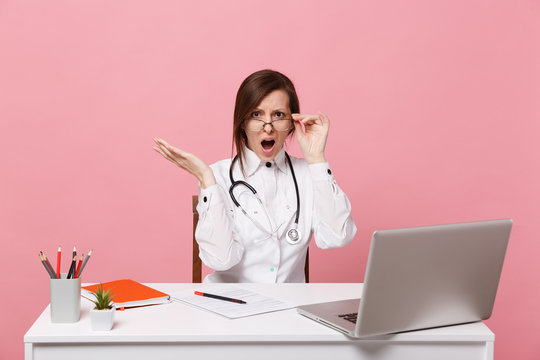 Tired Sad Female Doctor Sits At Desk Work On Computer With Medical Document In Hospital Isolated On Pastel Pink Wall Background. Woman In Medical Gown Glasses Stethoscope. Healthcare Medicine Concept.