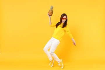 Fototapeta na wymiar Funny young woman in heart glasses dancing, holding fresh ripe pineapple fruit isolated on yellow orange wall background in studio. People vivid lifestyle, relax vacation concept. Mock up copy space.