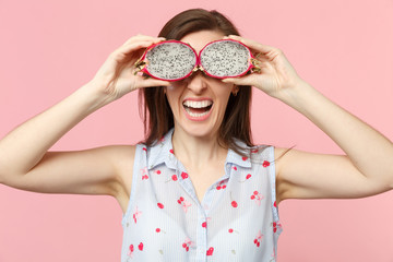 Laughing young woman holding, covering eyes with halfs of fresh ripe pitahaya dragon fruit isolated on pink pastel wall background. People vivid lifestyle relax vacation concept. Mock up copy space.