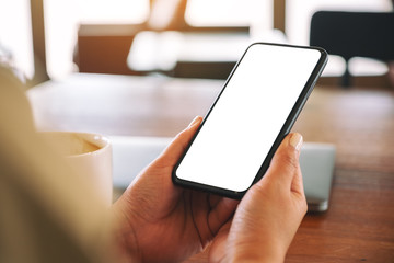 Mockup image of hands holding black mobile phone with blank desktop screen with laptop and coffee cup on the table