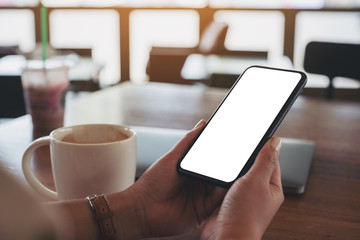Mockup image of hands holding black mobile phone with blank desktop screen with laptop and coffee cup on the table