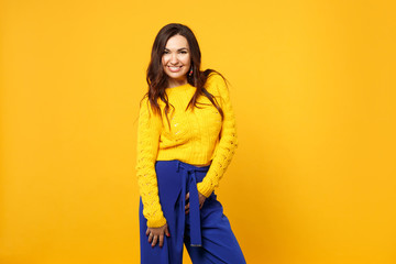 Portrait of smiling young woman in sweater, blue trousers standing, looking camera isolated on yellow orange wall background in studio. People sincere emotions, lifestyle concept. Mock up copy space.