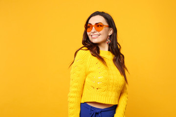 Portrait of pretty smiling young woman in sweater, blue trousers, heart glasses looking aside isolated on yellow orange wall background. People sincere emotions, lifestyle concept. Mock up copy space.