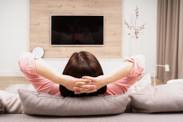 Back view of beautiful young woman resting in luxury appartments, sitting on the sofa and watching...