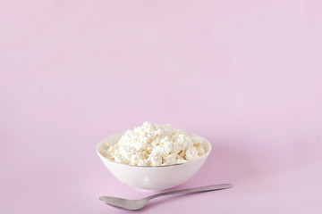 Fresh cottage cheese for breakfast in a white bowl and a spoon in the center of the frame on a pink background