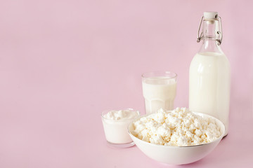Fresh cottage cheese for breakfast in a white bowl, sour cream, bottle of milk and glass of milk on a pink background