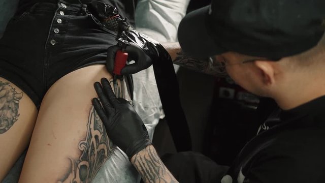 Master man tattoo in glasses draws the color paint on the clients tattoo. Tattoo artist holding tattoo machine in black sterile gloves