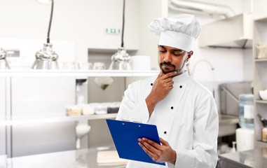 cooking, profession and people concept - male indian chef in toque reading menu on clipboard and thinking over restaurant kitchen background