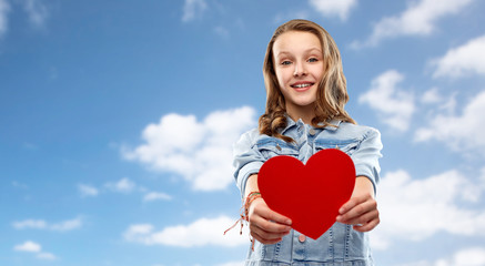 valentine's day, love and people concept - smiling pretty teenage girl in denim jacket holding red heart over blue sky and clouds background