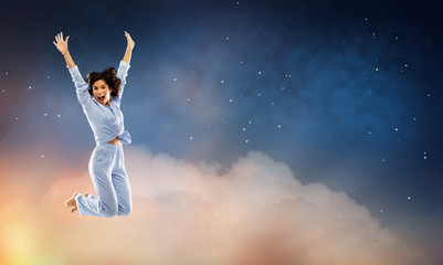 fun, people and bedtime concept - happy young woman full of energy in blue pajama jumping over starry night sky background