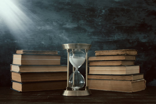 Hourglass As Time Passing Concept And Old Books In Front Of Black Wall Background. Conceptual Photo On History, Fantasy And Education