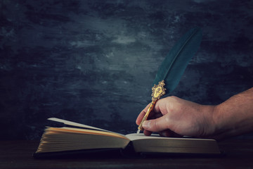 Old feather quill ink pen with inkwell and old books over wooden desk in front of black wall...