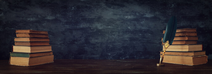 Old feather quill ink pen with inkwell and old books over wooden desk in front of black wall...
