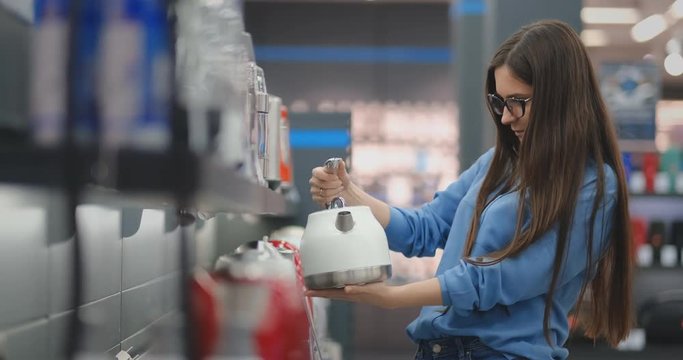 Counter of electronics store. Young beautiful brunette woman holding an electric kettle in her hands, studying the price tags, characteristics and design of the model