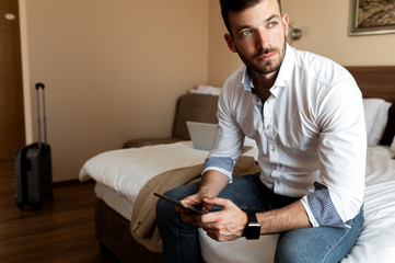 Young businessman working from hotel room on business trip, man sitting on bed and using tablet.