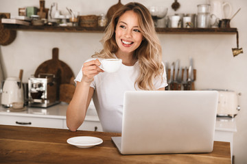 Image of cute blond woman drinking tea and using laptop while sitting at wooden table in apartment