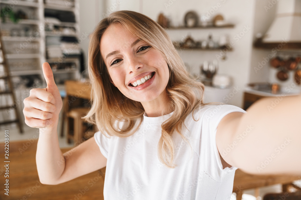 Wall mural Image closeup of cheery blond woman showing thumb up while taking selfie photo at home