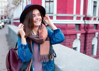 Fototapeta premium Outdoor lifestyle fashion portrait of young stylish hipster woman walking on street,wearing cute trendy outfit.Young woman with curly dark hair smiling to camera in city on city building background.
