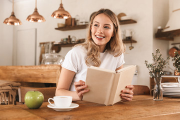 Portrait of smart blond woman reading book and drinking coffee while sitting in cozy cafe indoor