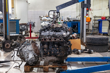 A detached engine suspended on a blue crane and a gear box on a lifting table in a vehicle repair shop. Auto service industry