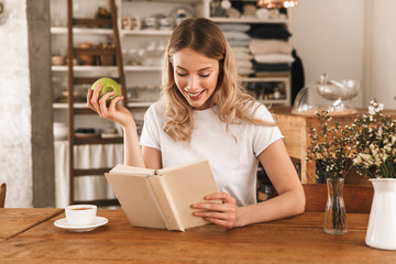 Portrait of charming blond woman reading book and eating green apple while sitting in cozy cafe indoor