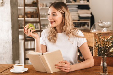 Portrait of smart blond woman reading book and eating green apple while sitting in cozy cafe indoor