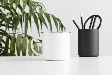 Enamel mug mockup with workspace accessories on a white table and a palm plant.